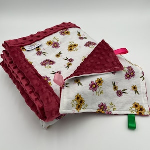 May include: A soft, white baby blanket with a floral print and a burgundy minky backing. The blanket has a pink ribbon and a green tag.