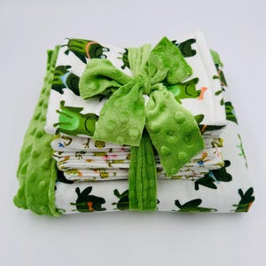 May include: A stack of baby blankets and washcloths tied with a green bow. The blankets feature a white background with green frog and floral patterns. A green, textured blanket is also included.