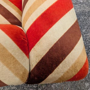 May include: Close-up of a section of a retro-style sofa or chair. The upholstery features a repeating pattern of diagonal stripes in shades of red, brown, beige, and cream. The fabric appears to be a soft, possibly velvet, material.