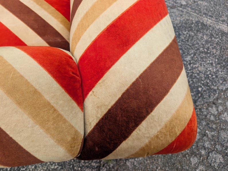 May include: Close-up of a section of a retro-style sofa or chair. The upholstery features a repeating pattern of diagonal stripes in shades of red, brown, beige, and cream. The fabric appears to be a soft, possibly velvet, material.