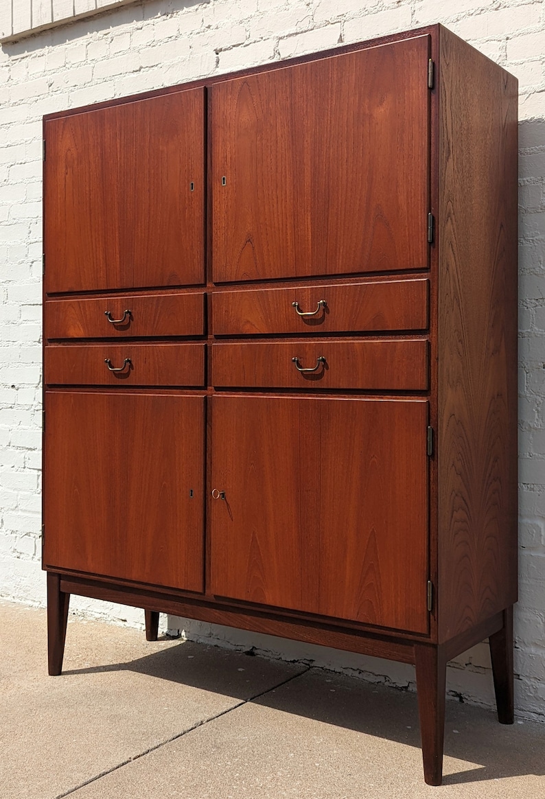 May include: A mid-century modern cabinet with two doors and four drawers. The cabinet is made of wood and has a rich brown finish. The cabinet has a keyhole on the right door and brass handles on the drawers.