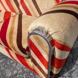 May include: Close-up of a vintage sofa with a striped pattern in shades of red, brown, and beige. The armrest features a curved, silver-colored metal frame. The fabric has a soft, textured appearance.