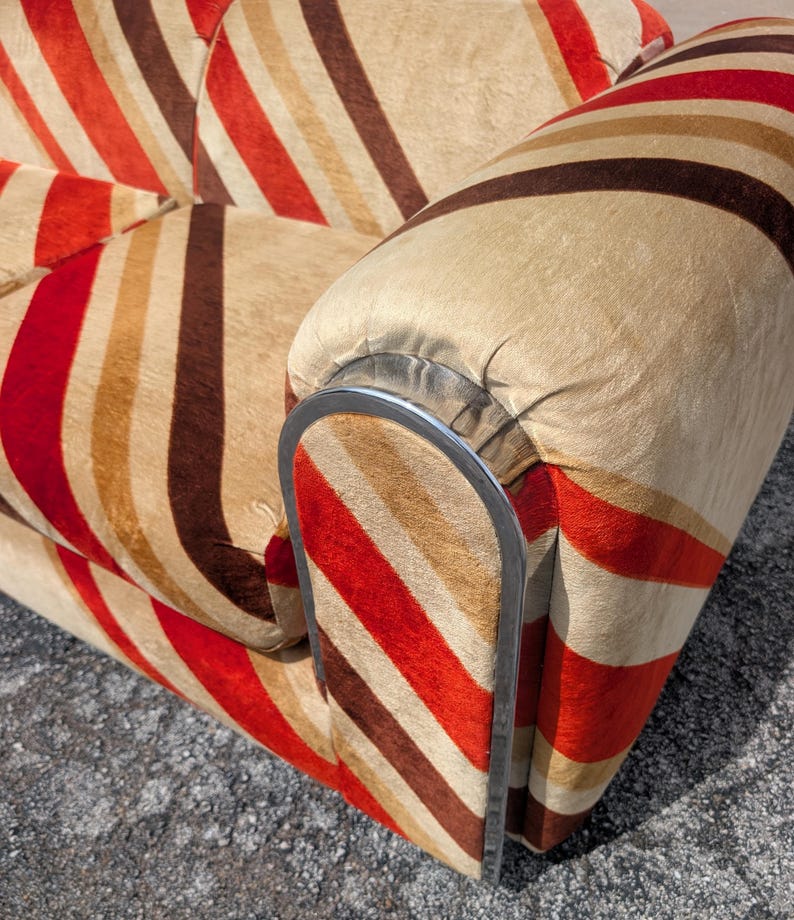 May include: Close-up of a vintage sofa with a striped pattern in shades of red, brown, and beige. The armrest features a curved, silver-colored metal frame. The fabric has a soft, textured appearance.