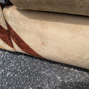 May include: Close-up of a section of a beige upholstered sofa with a brown and red striped pattern on the side. The sofa appears to be made of a soft, textured fabric. The background is a dark, textured surface.