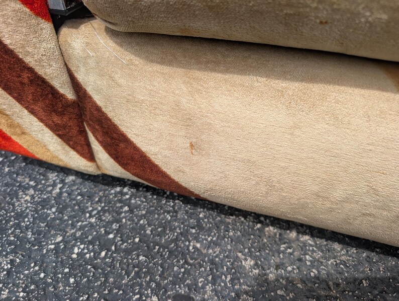 May include: Close-up of a section of a beige upholstered sofa with a brown and red striped pattern on the side. The sofa appears to be made of a soft, textured fabric. The background is a dark, textured surface.