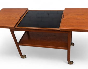 May include: A mid-century modern teak wood bar cart with a black center surface and two folding side leaves. The cart has a lower shelf and rolls on brass-toned casters. The wood has a warm, reddish-brown tone.