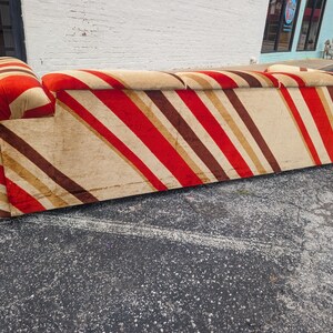 May include: A vintage sofa with a bold, striped pattern in red, brown, and beige. The sofa has a rounded armrest and a low profile. The fabric appears to be a soft, possibly velvet-like material, adding a touch of luxury to the design.