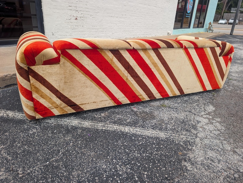 May include: A vintage sofa with a bold, striped pattern in red, brown, and beige. The sofa has a rounded armrest and a low profile. The fabric appears to be a soft, possibly velvet-like material, adding a touch of luxury to the design.