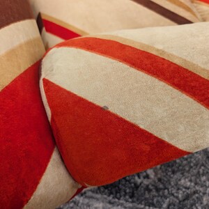 May include: Close-up of a section of a pillow with a retro design. The pillow features bold, diagonal stripes in shades of red, brown, tan, and cream. The fabric appears to be a soft, textured material, creating a vintage aesthetic.