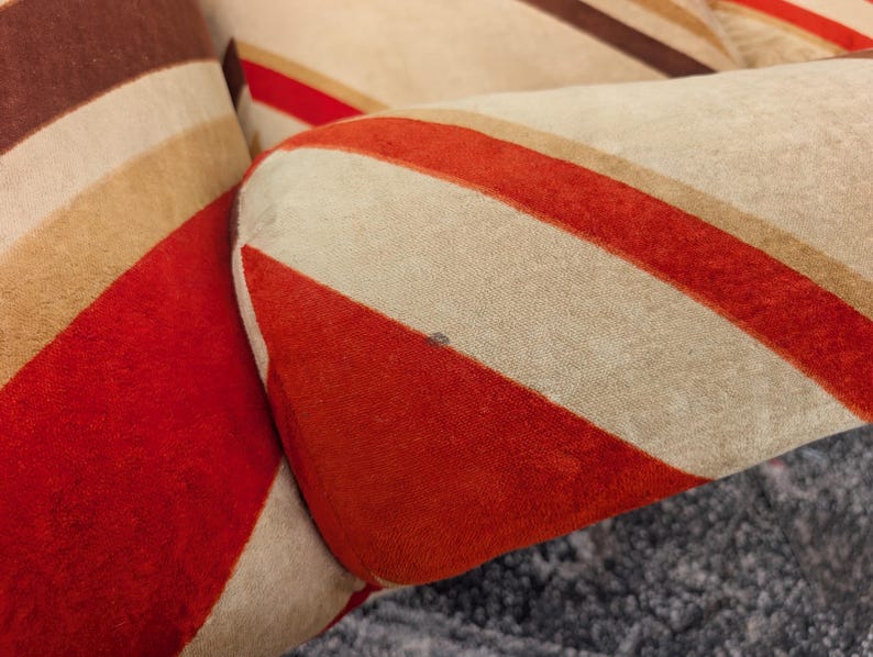 May include: Close-up of a section of a pillow with a retro design. The pillow features bold, diagonal stripes in shades of red, brown, tan, and cream. The fabric appears to be a soft, textured material, creating a vintage aesthetic.