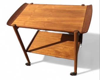 May include: A wooden serving cart with two shelves and wheels. The cart is made of light brown wood and has a simple, mid-century modern design.