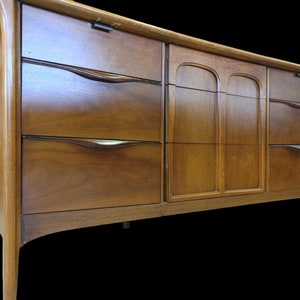 May include: A mid-century modern dresser with six drawers and two doors. The dresser is made of wood and has a warm brown finish. The drawers have curved metal handles.