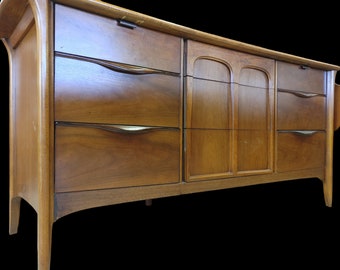 May include: A mid-century modern dresser with six drawers and two doors. The dresser is made of wood and has a warm brown finish. The drawers have curved metal handles.