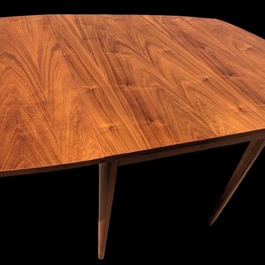 May include: A wooden dining table with a rectangular top and four legs. The table top is made of walnut wood and has a natural finish. The legs are tapered and have a dark brown finish.