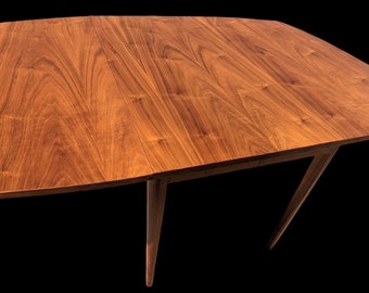 May include: A wooden dining table with a rectangular top and four legs. The table top is made of walnut wood and has a natural finish. The legs are tapered and have a dark brown finish.