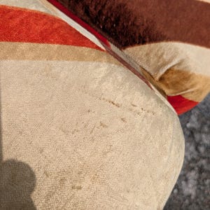 May include: Close-up of a section of a striped upholstered cushion. The stripes alternate between red, brown, and beige. The fabric has a soft, velvety texture, and the cushion appears to be well-worn.