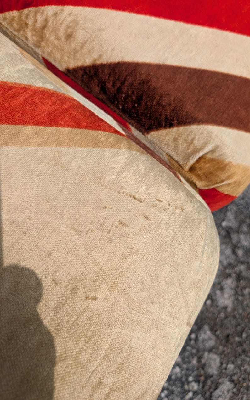 May include: Close-up of a section of a striped upholstered cushion. The stripes alternate between red, brown, and beige. The fabric has a soft, velvety texture, and the cushion appears to be well-worn.