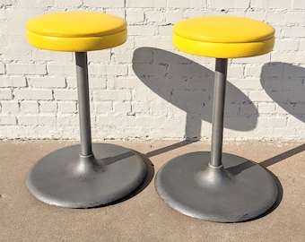 May include: Two yellow vinyl topped stools with gray metal bases. The stools are on a white brick background.