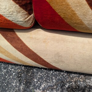 May include: Close-up of a section of a sofa with a retro design. The fabric features diagonal stripes in shades of orange, brown, and cream. The sofa's cushions are visible, showcasing the textured material and color pattern.