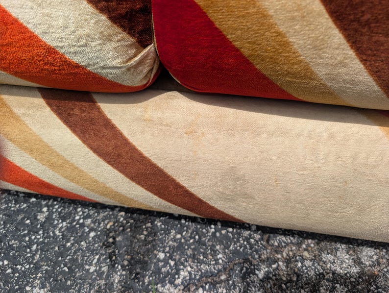 May include: Close-up of a section of a sofa with a retro design. The fabric features diagonal stripes in shades of orange, brown, and cream. The sofa's cushions are visible, showcasing the textured material and color pattern.