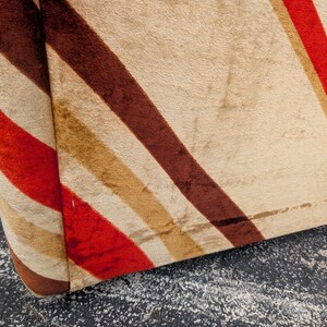 May include: Close-up of a section of a vintage upholstered chair. The fabric features diagonal stripes in red, brown, tan, and cream colors. The rounded edge of the chair is visible, showcasing the fabric's texture and pattern.
