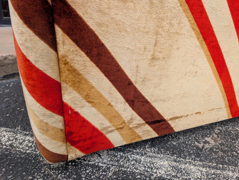 May include: Close-up of a section of a vintage upholstered chair. The fabric features diagonal stripes in red, brown, tan, and cream colors. The rounded edge of the chair is visible, showcasing the fabric's texture and pattern.