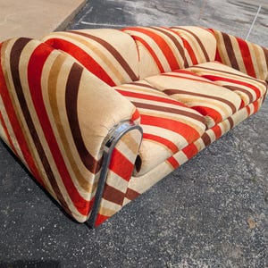May include: A vintage sofa with a bold striped pattern in shades of red, brown, and beige. The sofa features rounded arms and a chrome trim along the base. The cushions are also striped, creating a cohesive design.