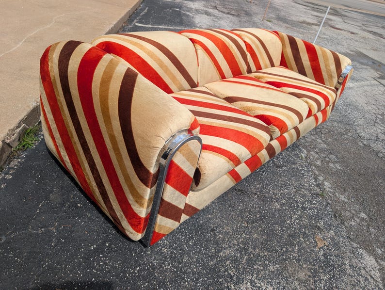 May include: A vintage sofa with a bold striped pattern in shades of red, brown, and beige. The sofa features rounded arms and a chrome trim along the base. The cushions are also striped, creating a cohesive design.