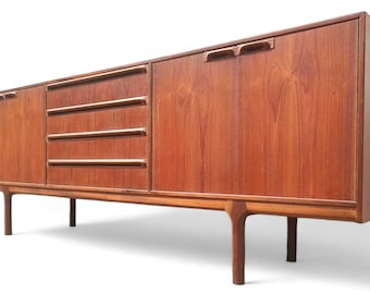 May include: A mid-century modern teak wood sideboard with three drawers and two cabinet doors. The sideboard is a rich brown colour and features simple, elegant hardware.