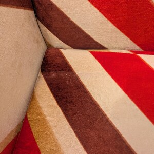 May include: Close-up of a section of a sofa with a bold striped pattern. The design features wide stripes in red, brown, and cream colors. The fabric appears to be soft and textured, with visible seams at the corner.