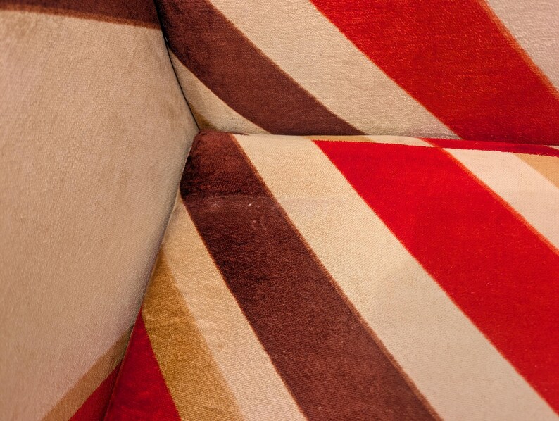 May include: Close-up of a section of a sofa with a bold striped pattern. The design features wide stripes in red, brown, and cream colors. The fabric appears to be soft and textured, with visible seams at the corner.