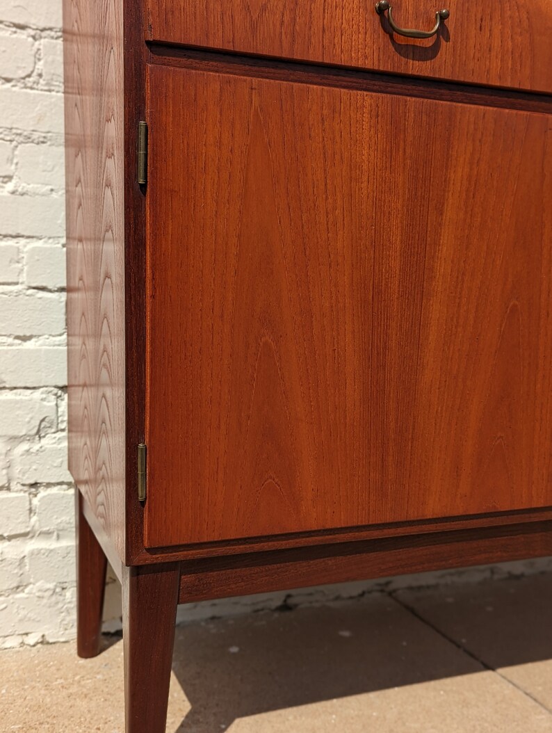 May include: A vintage wooden cabinet with a single door and a top drawer. The cabinet is made of dark wood with a rich grain pattern. The door has a brass handle and hinges. The cabinet is sitting on four legs.