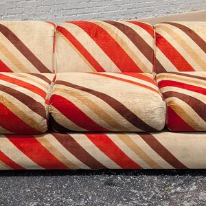 May include: A three-cushion sofa with a distinctive striped pattern in red, brown, and beige. The sofa has rounded arms and a low profile, offering a vintage aesthetic. The stripes run diagonally across the cushions and frame.