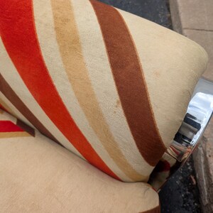 May include: Close-up of a vintage armchair with a striped pattern in shades of red, brown, and beige. The chair has a chrome armrest. The fabric has a textured appearance.