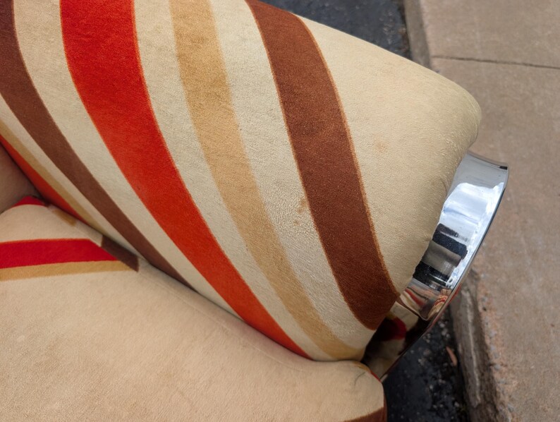 May include: Close-up of a vintage armchair with a striped pattern in shades of red, brown, and beige. The chair has a chrome armrest. The fabric has a textured appearance.