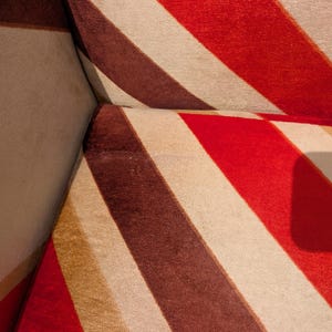 May include: Close-up of a section of a sofa with a bold, striped pattern. The stripes alternate between red, brown, beige, and tan. The fabric appears to be soft and textured, with a slight sheen.