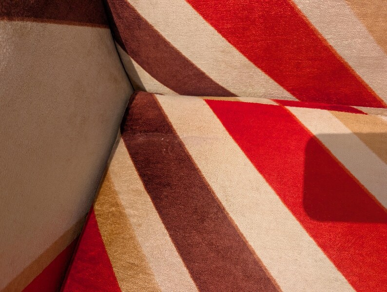 May include: Close-up of a section of a sofa with a bold, striped pattern. The stripes alternate between red, brown, beige, and tan. The fabric appears to be soft and textured, with a slight sheen.