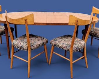 May include: A light brown wooden dining set consisting of an oval table and four chairs. Each chair features a curved backrest and a seat upholstered in a fabric with a geometric pattern in shades of gray, blue, and brown. The table has a smooth surface.