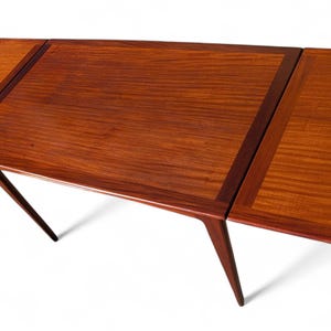 May include: A mid-century modern wooden table with a rectangular top and angled legs. The table is made of a rich, reddish-brown wood with a contrasting darker border. The table's leaves are extended, showcasing the wood grain.
