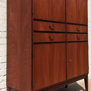 May include: A tall, wooden cabinet with two doors and three drawers. The cabinet is made of dark wood with a rich grain pattern. The doors have brass handles and the drawers have simple, metal pulls. The cabinet is standing on four legs.