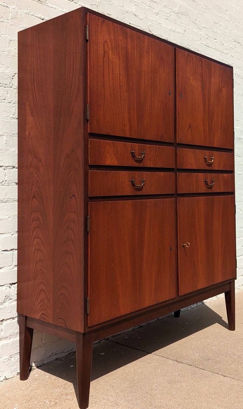 May include: A tall, wooden cabinet with two doors and three drawers. The cabinet is made of dark wood with a rich grain pattern. The doors have brass handles and the drawers have simple, metal pulls. The cabinet is standing on four legs.