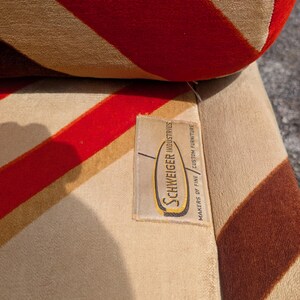 May include: Close-up of a vintage upholstered chair with a striped pattern in red, tan, and brown. The fabric has a soft, velvety texture. A tag reads "Schweiger Industries, Makers of Fine Custom Furniture."