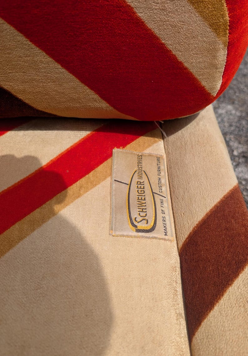 May include: Close-up of a vintage upholstered chair with a striped pattern in red, tan, and brown. The fabric has a soft, velvety texture. A tag reads "Schweiger Industries, Makers of Fine Custom Furniture."