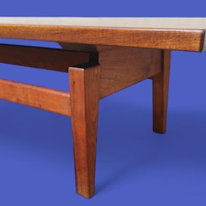 May include: A mid-century modern coffee table with a rectangular top and four tapered legs. The table is made of wood with a warm, reddish-brown finish. The tabletop has a lighter, natural wood color.