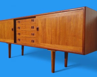 May include: A mid-century modern teak sideboard with four drawers and two doors. The sideboard is a warm honey brown colour and features brass hardware. The sideboard stands on four tapered legs.
