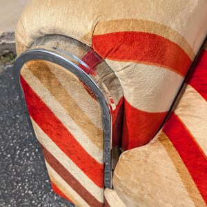 May include: Close-up of a vintage armchair with a striped pattern in red, brown, and beige. The chair has a curved chrome armrest. The fabric has a soft, textured appearance, suggesting a comfortable seating option.