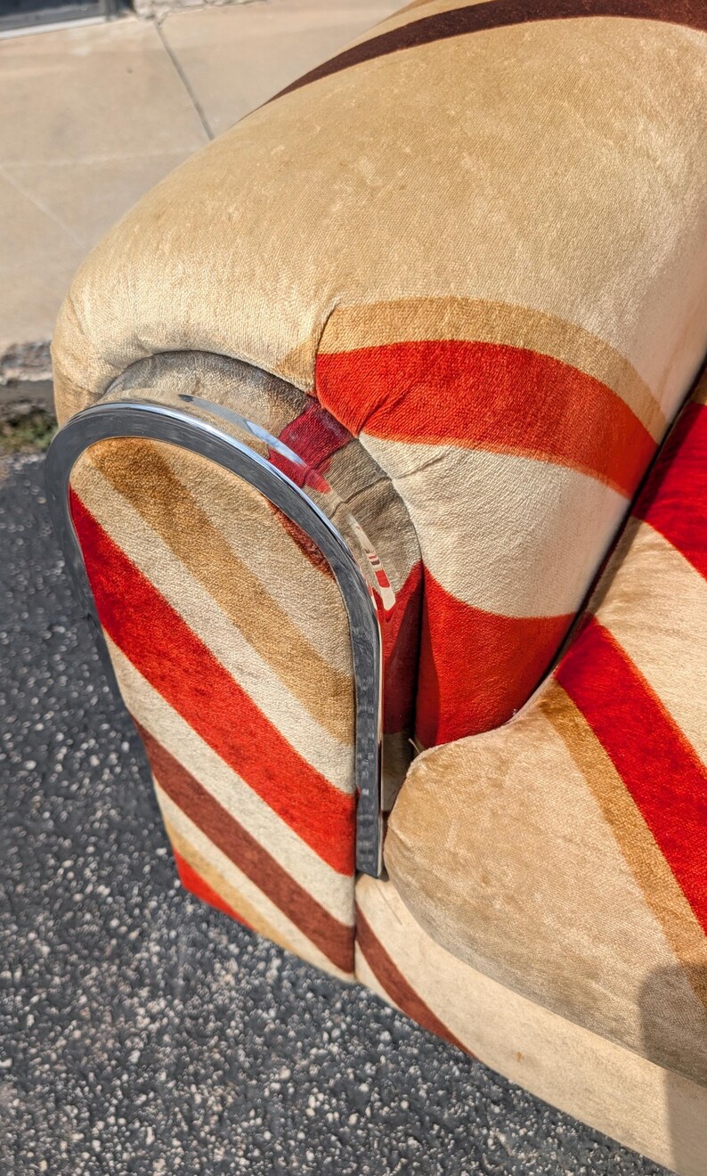 May include: Close-up of a vintage armchair with a striped pattern in red, brown, and beige. The chair has a curved chrome armrest. The fabric has a soft, textured appearance, suggesting a comfortable seating option.