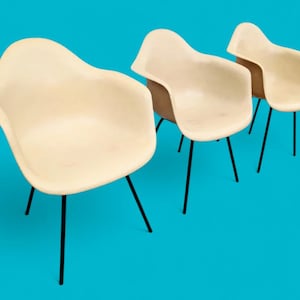 May include: Three cream-colored armchairs with black metal legs are arranged on a bright blue background. The chairs have a curved, organic shape and a mid-century modern aesthetic. The chairs are made of a smooth, solid material.
