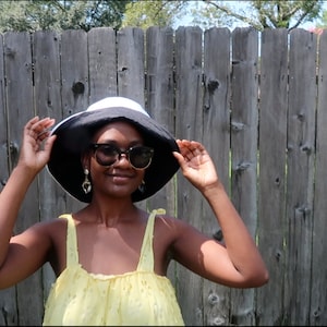 May include: A person wearing a black and white bucket hat, sunglasses, and a yellow sleeveless dress. The person is standing in front of a wooden fence.