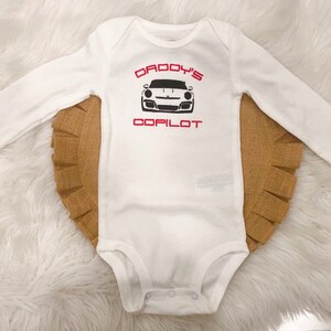 May include: White long-sleeved baby onesie with red text that says "Daddy's Copilot" and a black and white image of a sports car.